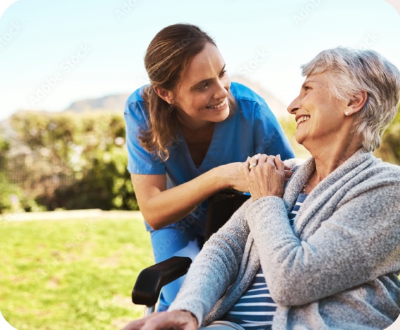 A caregiver in blue scrubs laughing with a resident in a wheelchair outdoors