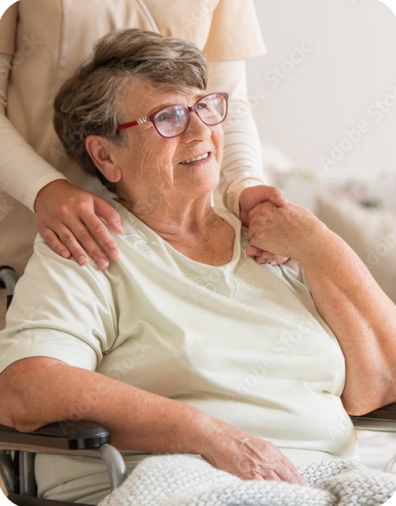 A smiling older woman in a wheelchair holding her caregiver&rsquo;s hand